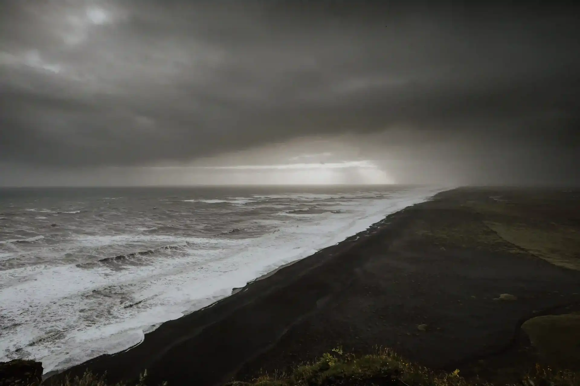 A bleak, dramatic view of a black sand beach with large waves under a dark, stormy sky. A ray of light breaks through the heavy clouds on the horizon.