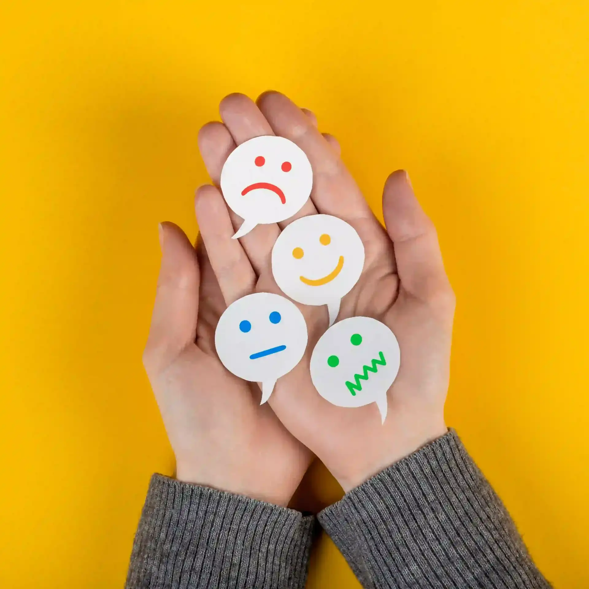 Top-down view of two hands cradling four paper cut-out speech bubbles on a bright yellow background. Each bubble features a different emoji-style face, representing various emotions.